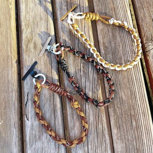 May include: Three braided bracelets in shades of brown, yellow, black, and white, with a toggle clasp. The bracelets are arranged on a wooden surface, showcasing their intricate woven design and colour variations. The bracelets are a stylish accessory.