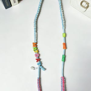 May include: A light blue and white braided strap with colorful accents. The strap features a series of rainbow-colored letter beads spelling out a word. Additional details include green, orange, and pink accents.
