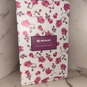 May include: A rectangular cardboard box featuring a white background with a repeating pattern of pink roses and green stems. A central rectangular label with a dark purple border displays the text STANLEY and LOVESHACKFANCY in white.