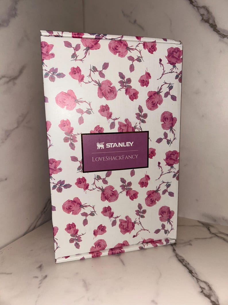 May include: A rectangular cardboard box featuring a white background with a repeating pattern of pink roses and green stems. A central rectangular label with a dark purple border displays the text STANLEY and LOVESHACKFANCY in white.
