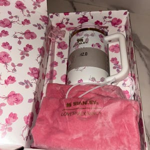 May include: A white Stanley H2.0 tumbler with a pink rose pattern, a white handle, and a matching straw, presented in a floral box with a pink velvet drawstring pouch featuring the embroidered text STANLEY and LOVESHACKFANCY.