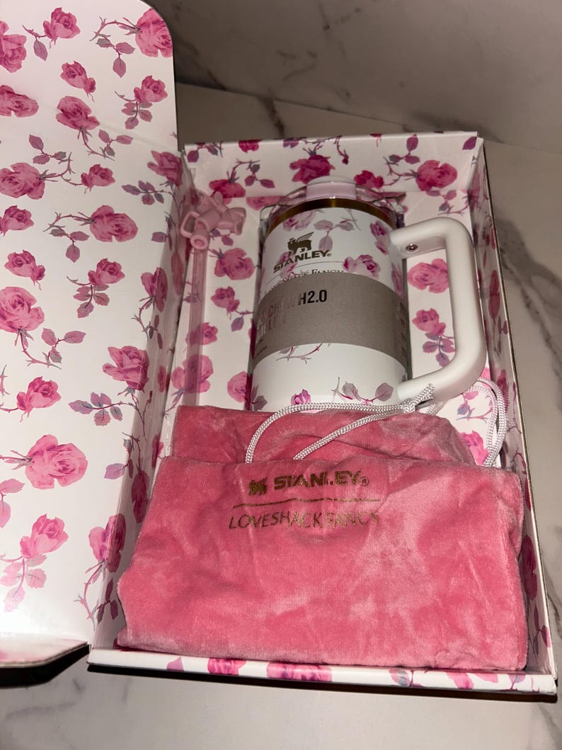 May include: A white Stanley H2.0 tumbler with a pink rose pattern, a white handle, and a matching straw, presented in a floral box with a pink velvet drawstring pouch featuring the embroidered text STANLEY and LOVESHACKFANCY.