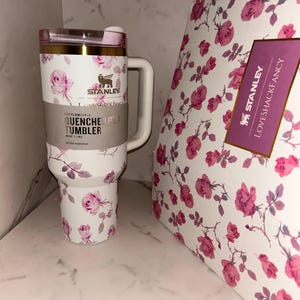 May include: A white Stanley Quencher H2.0 FlowState tumbler with a pink rose floral pattern, a gold rim, and a handle. The tumbler features a label reading Stanley, LoveShackFancy, and Quencher H2.0 Tumbler 40 oz 1.18 L. Beside it is a matching floral box.