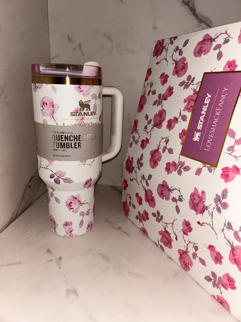 May include: A white Stanley Quencher H2.0 FlowState tumbler with a pink rose floral pattern, a gold rim, and a handle. The tumbler features a label reading Stanley, LoveShackFancy, and Quencher H2.0 Tumbler 40 oz 1.18 L. Beside it is a matching floral box.