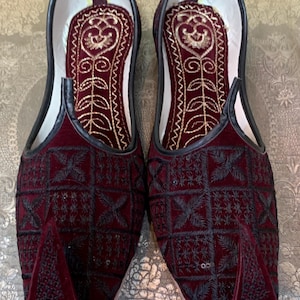 May include: A pair of burgundy velvet slippers with black embroidery and gold accents. The slippers have a pointed toe and a closed back.