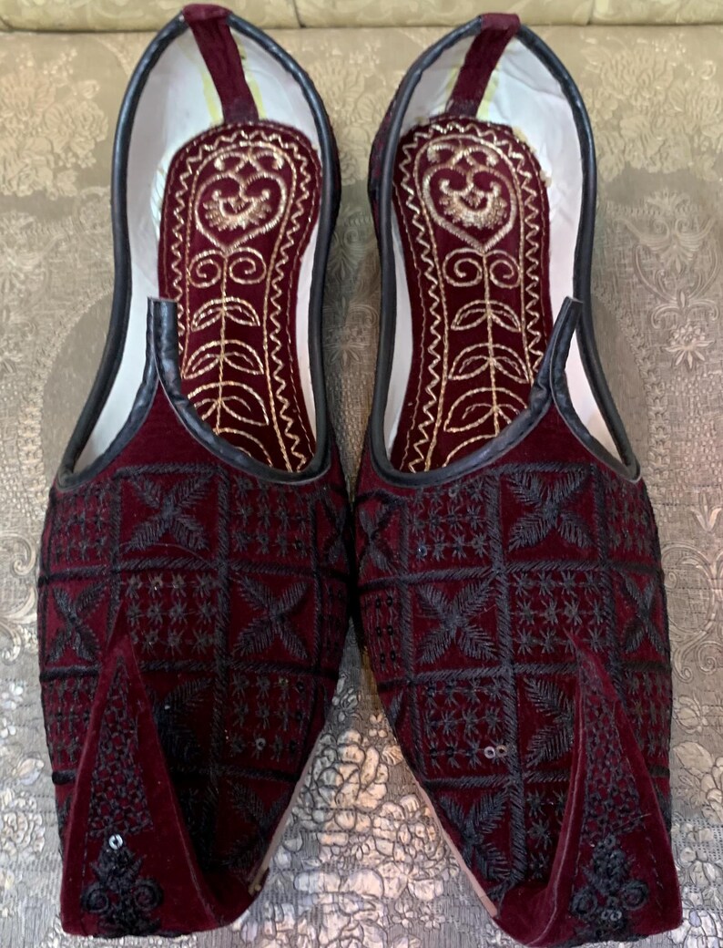 May include: A pair of burgundy velvet slippers with black embroidery and gold accents. The slippers have a pointed toe and a closed back.