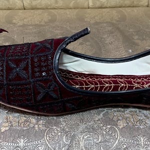 May include: A pair of burgundy velvet slippers with black embroidery and a pointed toe. The slippers have a white lining and a black trim.