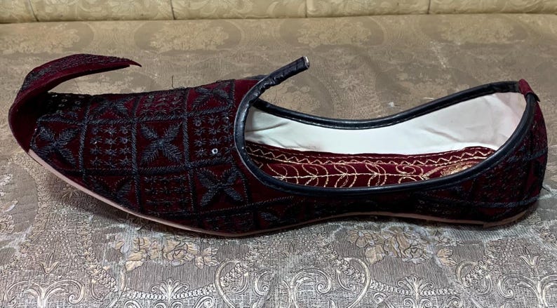 May include: A pair of burgundy velvet slippers with black embroidery and a pointed toe. The slippers have a white lining and a black trim.
