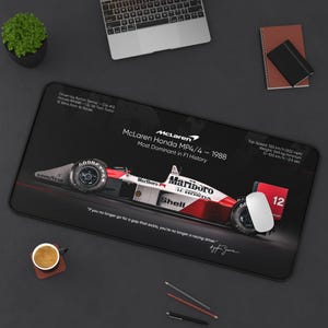 May include: Black rectangular mousepad featuring a McLaren Honda MP4/4 race car design. The design includes the car's specifications, the Marlboro and Shell logos, and a quote by Ayrton Senna. The mousepad also has a white mouse on it.
