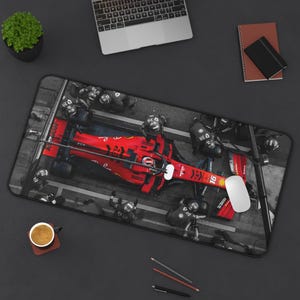 May include: A rectangular mousepad featuring a Formula 1 race car in red, with the number 16, surrounded by a pit crew in a black and white setting. A white mouse is on the right side of the mousepad.