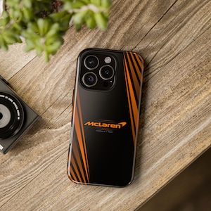 Könnte beinhalten: Schwarze Handyhülle mit orangefarbenen Streifen und dem McLaren Formula 1 Team Logo. Die Hülle liegt auf einer Holzoberfläche, mit einer Kamera und einer Pflanze im Hintergrund.