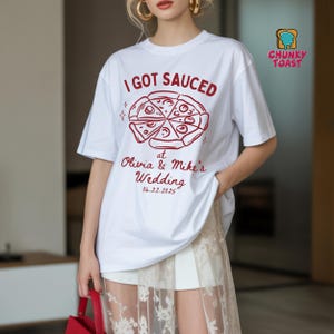 May include: White t-shirt with a red pizza graphic and the text "I GOT SAUCED" above "at Olivia & Mike's Wedding 06.22.2025". The shirt has short sleeves and a relaxed fit. A "Chunky Toast" logo is in the upper right corner.