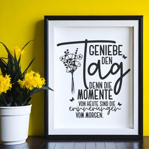 May include: A framed print with the German text "GENIE&szlig;E DEN Tag, DENN DIE MOMENTE VON HEUTE SIND DIE ERINNERUNGEN VOM MORGEN." The black and white design includes a floral bouquet and butterflies. A potted plant with yellow flowers sits to the left.