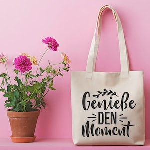 May include: A cream-colored tote bag with the German phrase "Genie&szlig;e DEN Moment" printed in black. The bag has long handles and is set against a pink background. A potted plant with pink and yellow flowers is on the left.