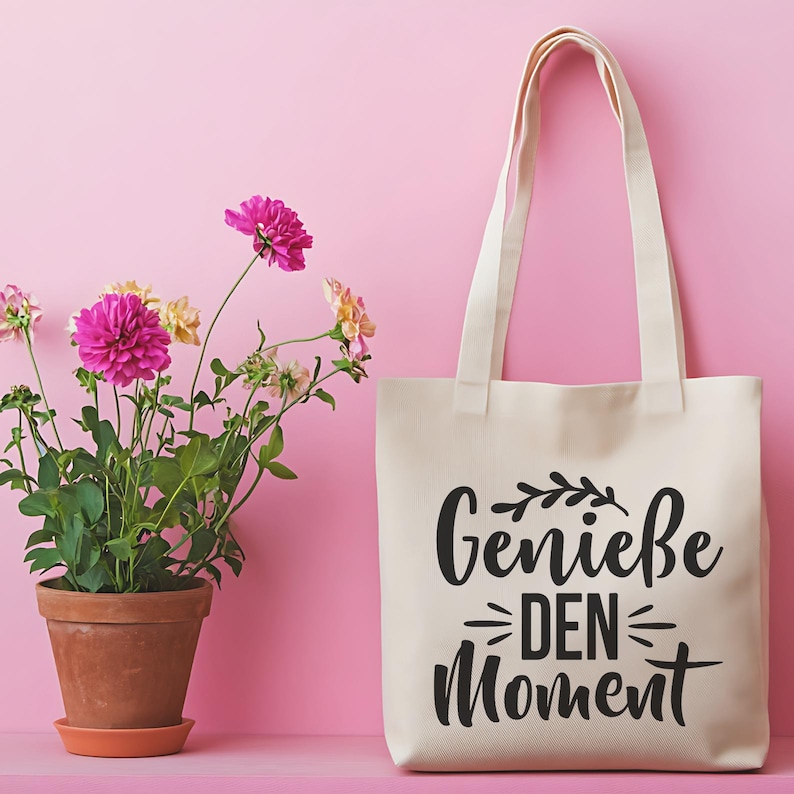 May include: A cream-colored tote bag with the German phrase "Genie&szlig;e DEN Moment" printed in black. The bag has long handles and is set against a pink background. A potted plant with pink and yellow flowers is on the left.