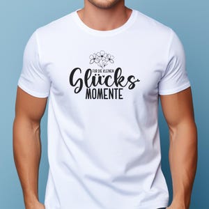 May include: White t-shirt with a black graphic design. The design includes the words "F&uuml;r die kleinen Gl&uuml;cks Momente" in a stylized font, along with a small floral illustration above the word "Gl&uuml;cks".