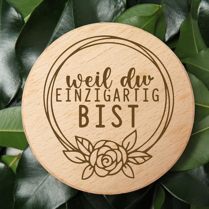 May include: A round wooden coaster with the German text "weil du EINZIGARTIG BIST" (because you are unique) in brown. The coaster features a floral design and is set against a background of green leaves.