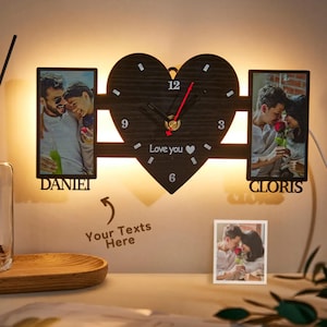 May include: A heart-shaped wall clock with a black face and a red second hand. It features two photo frames and the words "Love you." The clock has the names "DANIEL" and "CLORIS" below the photo frames.