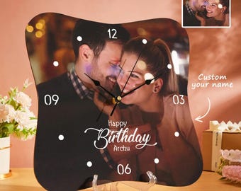 May include: A personalized clock with a photo of a couple, featuring the text "Happy Birthday Archu." The clock has a black background with white hour markers and hands. The clock is displayed on a clear stand.
