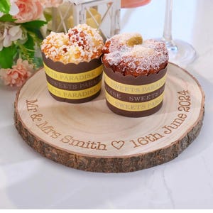 May include: Two cupcakes in decorative wrappers with the text "SWEETS PARADISE" are presented on a wooden serving board. The board is engraved with "Mr & Mrs Smith 16th June 2024". The cupcakes have different toppings.