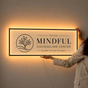 May include: A rectangular, backlit wall sign with a black frame and a white face. It features a black line-art tree logo, the text "EST 2024", "MINDFUL COUNSELING CENTER", and the tagline "Your Mind. Your Path. Our Support." in black lettering.