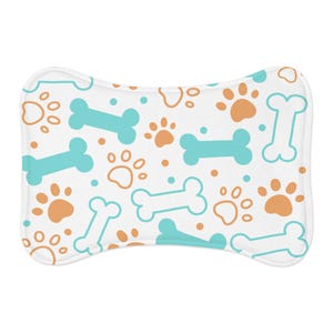 Puede incluir: Cama para mascotas con forma de hueso, con un fondo blanco y un patrón repetido de huesos azul claro, huellas de patas naranjas y pequeños puntos. La cama tiene un borde blanco.