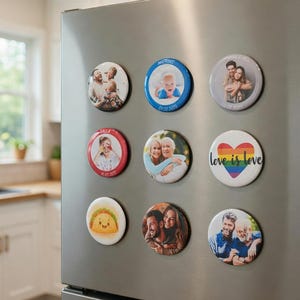 May include: A collection of nine circular magnets arranged in a grid on a stainless steel refrigerator. The magnets feature various designs including personal photographs of people, a cartoon taco, and a rainbow heart with the text love is love.