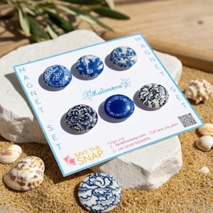 Könnte beinhalten: Ein Set dekorativer Magnete mit blau-weißen floralen Designs. Das Magnetset enthält sieben runde Magnete mit verschiedenen Mustern, darunter eine durchgehend blaue Mitte und florale Muster. Die Magnete werden auf einer weißen Karte mit dem Text "MAGNET SET" und "Kalimera" präsentiert.