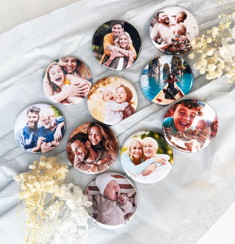 May include: A collection of nine circular photo buttons featuring various colour portraits of families, couples, and individuals, including a baby in a white hat, a person in a boat on blue water, and a child playing in sand, arranged on white paper.