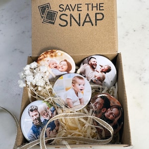 May include: A brown cardboard box with the text SAVE THE SNAP printed on the inside lid. Inside are four circular photo magnets featuring various family portraits, nestled in light-coloured paper filler with a small sprig of dried white flowers.