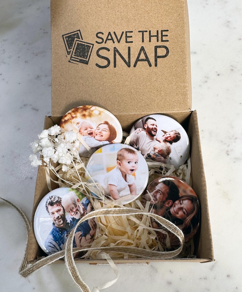 May include: A brown cardboard box with the text SAVE THE SNAP printed on the inside lid. Inside are four circular photo magnets featuring various family portraits, nestled in light-coloured paper filler with a small sprig of dried white flowers.