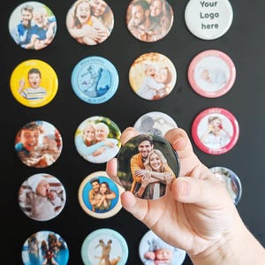 May include: A collection of circular pin-back buttons featuring various printed photographs of people, infants, and landscapes, with one button displaying the text Your Logo here, held in a hand against a plain dark background.