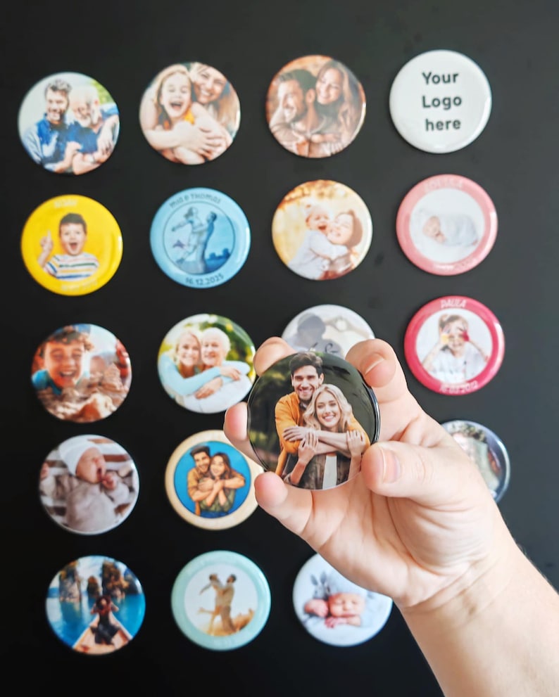 May include: A collection of circular pin-back buttons featuring various printed photographs of people, infants, and landscapes, with one button displaying the text Your Logo here, held in a hand against a plain dark background.