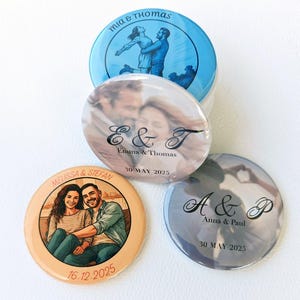 May include: Four circular pin-back buttons featuring personalised designs. One shows a blue illustration of a couple with the text Mia & Thomas. Another displays a photo of a couple with E & T, Emma & Thomas, and 30 May 2025. A third shows a couple with Melissa & Stefan and 16.12.2025. The last shows A & P, Anna & Paul, and 30 May 2025.
