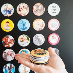 May include: A collection of circular custom photo magnets featuring various portraits and images, including one white magnet with the text Your Logo here. The magnets are displayed on a dark surface, with a stack of five magnets held in the foreground.