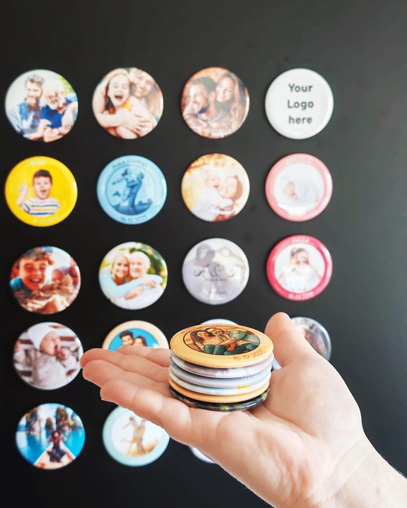 May include: A collection of circular custom photo magnets featuring various portraits and images, including one white magnet with the text Your Logo here. The magnets are displayed on a dark surface, with a stack of five magnets held in the foreground.
