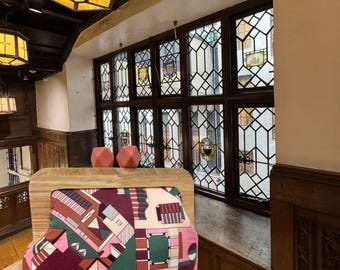May include: A wooden-framed clutch with a colorful geometric pattern in shades of pink, burgundy, and green. Two peach-colored geometric shapes sit atop the frame. The clutch is held in front of a window with stained glass.
