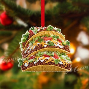 Pode incluir: Um enfeite de Natal em forma de três tacos empilhados. O enfeite tem uma fita vermelha para pendurar e é decorado com alface, tomate, queijo e carne. O enfeite está em um fundo desfocado de árvore de Natal.