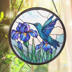 May include: A round stained glass window panel featuring a blue hummingbird and iris flowers. The hummingbird is depicted with detailed blue and turquoise feathers, hovering near the purple and blue iris blossoms. The panel is framed in a dark metal and hangs from a chain.