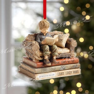 Puede incluir: Adorno decorativo que representa a una persona joven leyendo un libro, sentado en una silla en miniatura encima de una pila de libros. El adorno tiene una cinta roja para colgar. Los libros tienen texto en los lomos.