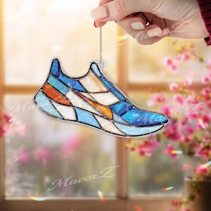 Puede incluir: Un atrapasoles de vidrieras con forma de zapatilla deportiva, que presenta un patrón geométrico de segmentos de vidrio azul, naranja y blanco unidos por líneas de soldadura de metal oscuro, suspendido de una fina cadena de metal plateado.