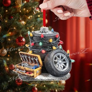 Könnte beinhalten: Ein Weihnachtsornament mit einem Stapel Reifen, der mit bunten Lichterketten geschmückt ist. Eine Werkzeugkiste mit Werkzeugen und ein Reifen mit silberner Felge sind ebenfalls enthalten. Das Ornament wird an einem roten Band gehalten.