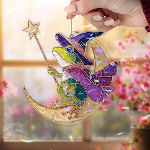 May include: A stained glass frog wearing a witch hat and cape, sitting on a crescent moon. The frog holds a star-topped wand. The artwork features green, purple, and gold colors, with a chain for hanging.