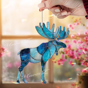 May include: A stained glass moose ornament featuring various shades of blue and teal glass pieces joined by dark metal solder lines. The moose is suspended by a thin silver chain and displays a textured, semi-translucent finish throughout.