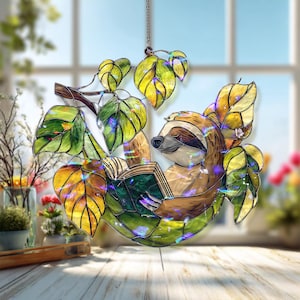 May include: Stained glass sloth reading a book, suspended from a chain. The sloth is brown and tan, with closed eyes, surrounded by green and yellow leaves. The leaves and sloth are lit with small, colourful lights, creating a whimsical, decorative piece.
