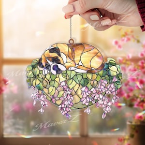 Puede incluir: Atrapasoles de vidriera con un gato durmiendo en un lecho de hojas verdes y flores de glicina rosa. El gato tiene pelaje marrón y blanco. El atrapasoles se sujeta con una cadena.