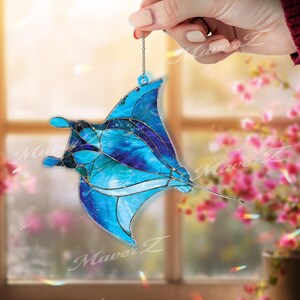 Puede incluir: Adorno de vidriera con forma de manta raya en tonos azules y morados, con una cadena plateada para colgar. El adorno tiene un diseño detallado con un patrón celestial y está sujeto por una mano.