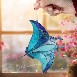 Puede incluir: Un atrapasoles de vidrieras con forma de mantarraya, que presenta varias piezas de vidrio en tonos azules y turquesas unidas por líneas de soldadura de metal oscuro. La pieza está suspendida por una fina cadena de metal plateado.