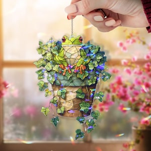 May include: A stained-glass frog ornament in a flower pot with ivy. The frog is green with red eyes and orange feet. The flower pot is brown and tan. The ivy is green with blue lights. The ornament is hanging from a silver chain.