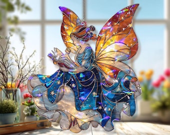 Op de afbeelding: Een gebrandschilderd glazen feeënornament met iriserende oranje en gele vleugels, een blauwe jurk en een boek. De fee hangt aan een ketting. Het ornament staat op een houten oppervlak met een vage achtergrond van bloemen en een raam.